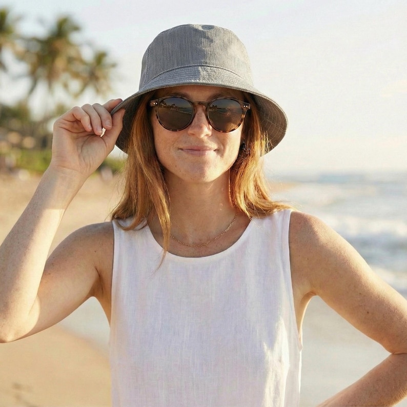 May include: A woman wearing a gray bucket hat and tortoiseshell sunglasses. She is wearing a white sleeveless top and a gold necklace. The background shows a beach with palm trees and the ocean.