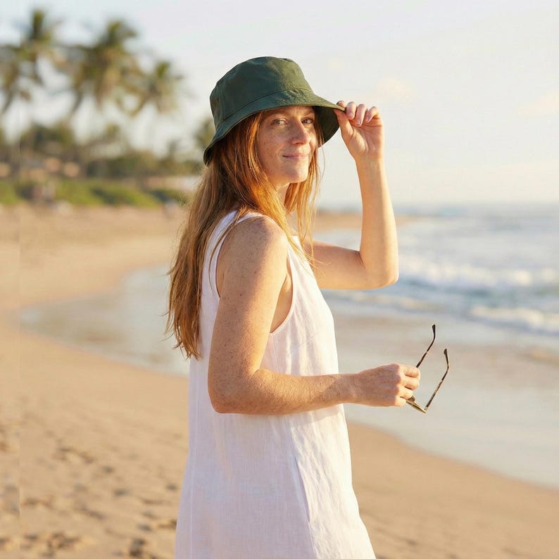 May include: A woman wearing a dark green bucket hat and a white sleeveless dress stands on a sandy beach. She holds sunglasses in her hand and smiles at the camera. The background includes the ocean and palm trees.