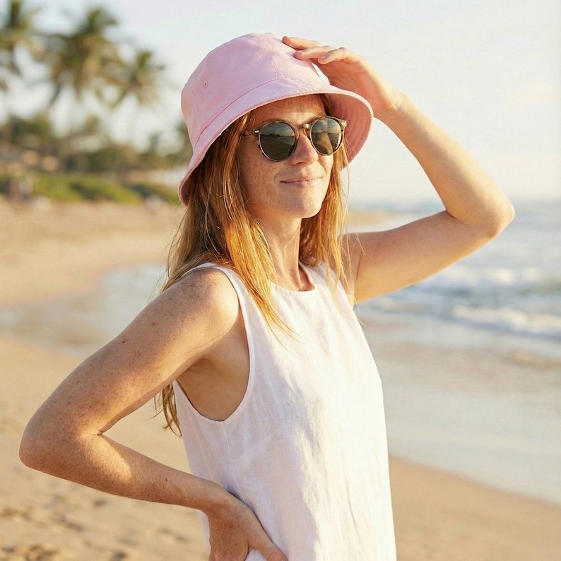 May include: A woman wearing a light pink bucket hat and sunglasses, standing on a beach. She is wearing a white sleeveless dress. The background shows a sandy beach and the ocean.