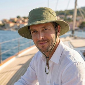 Peut inclure: Chapeau de pêcheur vert olive avec un large bord et une mentonnière réglable. Le chapeau est doté d'œillets d'aération et est fabriqué dans un tissu léger. Une personne porte le chapeau avec une chemise blanche.