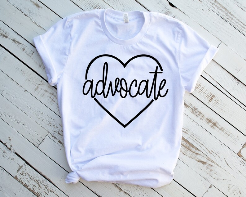 Puede incluir: Camiseta blanca con la palabra "advocate" en un dise&ntilde;o en forma de coraz&oacute;n negro. El texto est&aacute; en cursiva. La camiseta est&aacute; hecha de un material suave y tiene mangas cortas.
