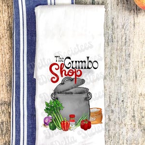 Könnte beinhalten: Weißes Küchentuch mit einer Grafik einer Kochtopf, Gemüse und dem Text "The Gumbo Shop" in Schwarz und Rot. Ein blau-weiß gestreiftes Handtuch ist links zu sehen. Das Bild enthält eine rote Paprika, Sellerie und eine kleine Flasche.
