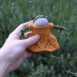 May include: A knitted frog wearing a bright orange dress. The frog is green and has black eyes. The frog is being held in a hand.