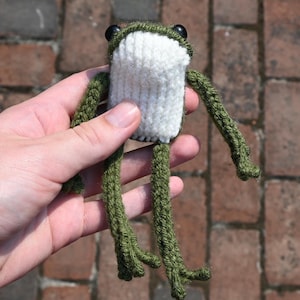 May include: A green knitted frog with white yarn for the belly and black button eyes. The frog is held in a hand against a brick background.