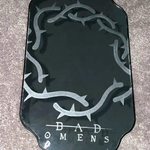 Puede incluir: Espejo decorativo negro con una forma única y un diseño de espinas plateadas en los bordes. Las palabras "BAD OMENS" están impresas en plata en la parte inferior. Tiene un marco de metal negro y una cadena para colgar.