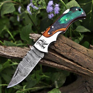 May include: A folding knife with a Damascus steel blade and a handle made of wood and green material. The handle features white dot accents, and the blade is engraved with the name "ROBERT" and the letter "R". The knife rests on a piece of wood.