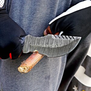 Puede incluir: Primer plano de una persona sosteniendo un cuchillo de acero Damasco con mango de madera. El cuchillo tiene una hoja texturizada y una punta afilada.