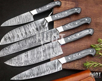 Puede incluir: Juego de cinco cuchillos de cocina de acero de Damasco con mangos de vetas de madera gris y virolas plateadas. Las hojas tienen un patrón único similar al agua. Los cuchillos están sobre una tabla de madera.