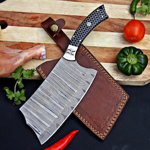 May include: A cleaver knife with a Damascus steel blade featuring a wavy pattern and a black handle with a white honeycomb design. The knife rests on a brown leather sheath with visible stitching and a metal snap button closure.
