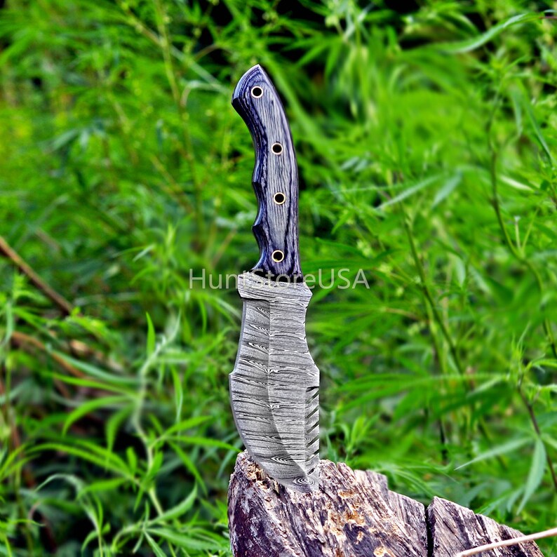 Puede incluir: Un cuchillo de caza de acero Damasco con mango de madera negra. El cuchillo est&aacute; apoyado en un toc&oacute;n de &aacute;rbol en una zona verde cubierta de hierba.