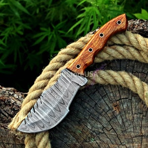 Puede incluir: Un cuchillo de caza de acero Damasco con mango de madera. La hoja tiene un patr&oacute;n ondulado y el mango es de color marr&oacute;n rojizo.