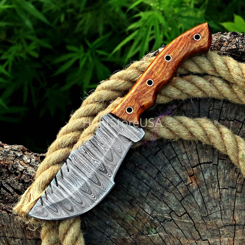 Puede incluir: Un cuchillo de caza de acero Damasco con mango de madera. La hoja tiene un patr&oacute;n ondulado y el mango es de color marr&oacute;n rojizo.