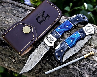 May include: Two folding pocket knives with blue and black wood handles and Damascus steel blades, one open and one closed. The open knife features the name Robert, while the closed one shows Emily Smith. Includes a brown leather sheath and a sharpener.