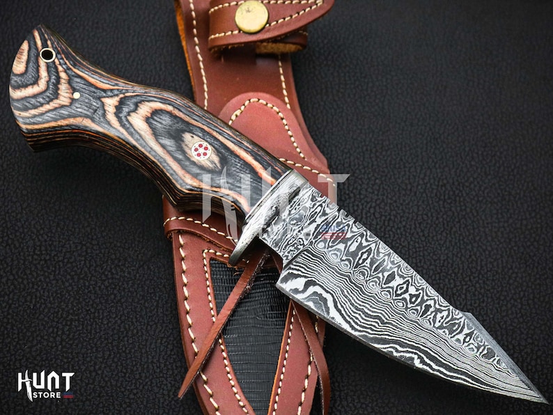 May include: A black and brown wood handle knife with a Damascus steel blade. The knife is in a brown leather sheath.