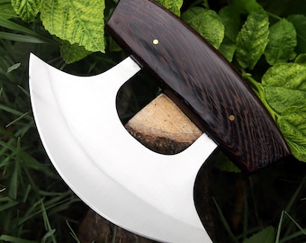 May include: A stainless steel mezzaluna knife with a curved blade and a dark wood handle. The handle is secured with brass rivets. The knife is designed for chopping herbs and vegetables. The knife is set against a backdrop of green leaves and grass.