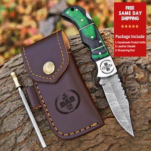 May include: A green and black pocket knife with a Damascus steel blade and a leather sheath. The knife has a wood handle and is engraved with the words "Best Dad Ever".