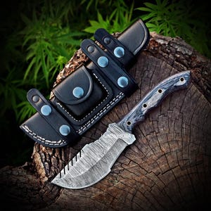 Puede incluir: Una funda de cuero negra con un cuchillo con una hoja estampada y un mango de madera negro y gris. El cuchillo est&aacute; sobre una superficie de madera.
