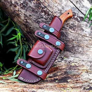 Puede incluir: Una funda de cuchillo de cuero marr&oacute;n con un cuchillo de mango de madera. La funda tiene m&uacute;ltiples correas con broches de metal azul y un bolsillo peque&ntilde;o.