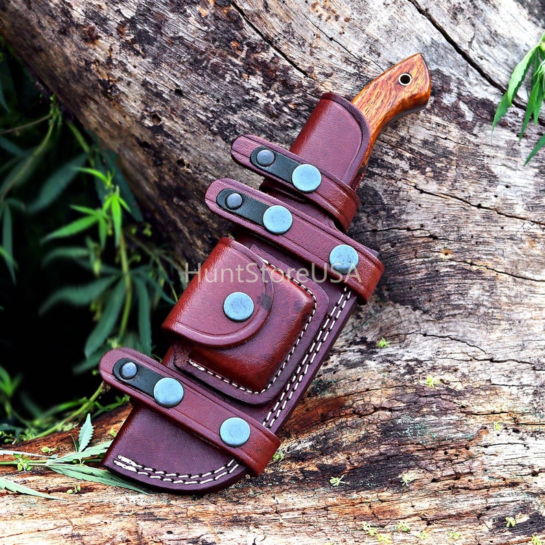 Puede incluir: Una funda de cuchillo de cuero marr&oacute;n con un cuchillo de mango de madera. La funda tiene m&uacute;ltiples correas con broches de metal azul y un bolsillo peque&ntilde;o.