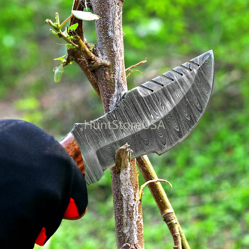 Puede incluir: Primer plano de un cuchillo de acero Damasco con mango de madera, que se utiliza para cortar una rama peque&ntilde;a. El cuchillo tiene un patr&oacute;n distintivo en la hoja y una punta afilada.