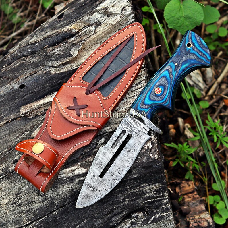May include: A blue and black patterned knife with a Damascus steel blade and a leather sheath. The knife has a decorative handle and a finger guard. The sheath has a belt loop and a snap closure. HuntStoreUSA is printed on the sheath.