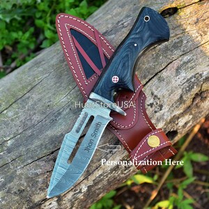 May include: A black and silver hunting knife with a wooden handle and a leather sheath. The knife has a personalized inscription that reads "John Smith".