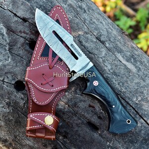 May include: A hunting knife with a black and grey handle and a leather sheath. The blade is made of Damascus steel and has a decorative pattern.