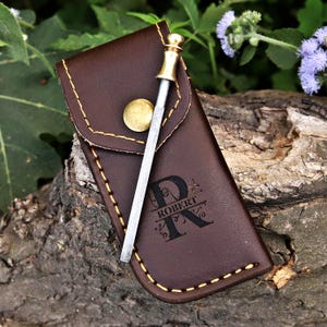 May include: A brown leather knife sharpener case with gold stitching and a gold button closure. A metal sharpening rod with a gold handle is inserted into the case. The case is personalized with the name "ROBERT" and a decorative initial "R".