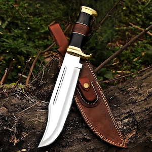 May include: A large hunting knife with a polished steel blade and a black, brown, and gold handle. The knife rests on a log with its brown leather sheath. The sheath has a decorative stitched pattern and a snap closure.