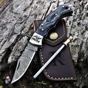 May include: A folding pocket knife with a Damascus steel blade and a dark wood handle featuring the engraved text ROBERT. The knife is paired with a brown leather sheath and a metal sharpening rod, both displaying the name ROBERT.