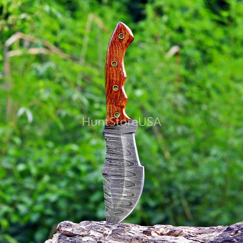 Puede incluir: Un cuchillo de caza con una hoja de acero Damasco y un mango de madera con detalles de lat&oacute;n. El cuchillo est&aacute; de pie sobre un trozo de madera.