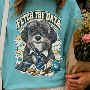 May include: A light blue t-shirt featuring a graphic of a fluffy dog wearing glasses and a necktie, surrounded by office supplies and the text "FETCH THE DATA". The shirt is paired with a white baseball cap and a white tiered skirt.