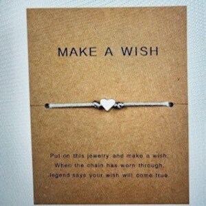 May include: White string bracelet with a white heart charm. The card reads "MAKE A WISH" and "Put on this jewellery and make a wish, When the chain has worn through, legend says your wish will come true."