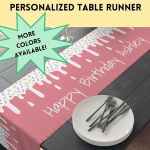 May include: A personalized table runner with a pink design resembling dripping frosting and colorful sprinkles. The text "Happy Birthday Ashley" is displayed in white. The words "More Colors Available!" are in a green starburst.