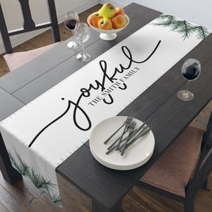 May include: White table runner with the word "joyful" in a black script font, along with "THE SMITH FAMILY". The runner is decorated with green pine branches and is set on a dark wood table with a bowl of apples and wine glasses.
