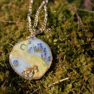 May include: A gold chain necklace with a round pendant featuring a crescent moon, stars, and pressed purple flowers encased in clear resin.