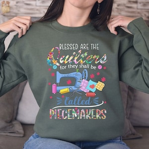 May include: Green sweatshirt with a sewing machine and colorful fabric scraps. The text reads "Blessed are the Quilters for they shall be called Peacemakers".