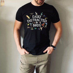 May include: Black t-shirt with the text "EASILY DISTRACTED BY BIRDS" in white, surrounded by colorful bird illustrations. The shirt is short-sleeved and made of a soft material. The shirt is worn by a person.