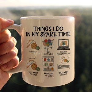 May include: White ceramic mug with a black outline of a person crocheting and the text "Things I do in my spare time". The mug has six illustrations of activities related to crocheting, including crocheting, buying yarn, researching crochet patterns, talking about crochet, rearranging yarn, and dreaming about crochet.