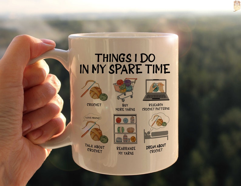 May include: White ceramic mug with a black outline of a person crocheting and the text "Things I do in my spare time". The mug has six illustrations of activities related to crocheting, including crocheting, buying yarn, researching crochet patterns, talking about crochet, rearranging yarn, and dreaming about crochet.