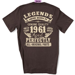 Op de afbeelding: Een donkerbruin T-shirt met een vintage-stijl grafisch ontwerp met de tekst "Legends were born in 1961 Aged Perfectly All Original Parts".