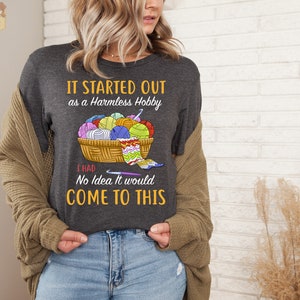 May include: Dark gray t-shirt with the text "IT STARTED OUT as a Harmless Hobby I HAD No Idea It would COME TO THIS." The shirt features a graphic of a basket filled with yarn balls and crochet needles. The shirt is a crew neck.