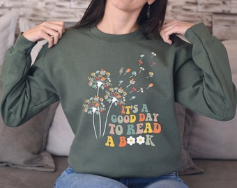 May include: A green sweatshirt featuring a dandelion design made of books. The dandelion seeds are blowing away. The text "It's a good day to read a book" is printed in rainbow colours on the sweatshirt.