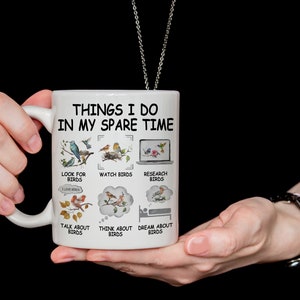 May include: White ceramic mug with a black outline and the text "Things I do in my spare time" with a list of bird-related activities. The mug is being held by a person's hands.
