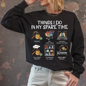 May include: Black sweatshirt with a white graphic that says "Things I do in my spare time" and shows a series of illustrations related to crocheting, including yarn, crochet hooks, and a shopping cart.