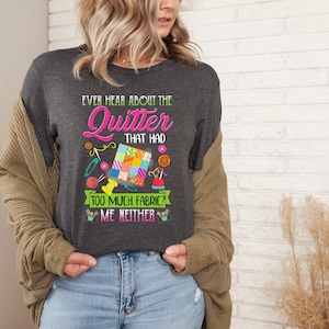 May include: A dark gray t-shirt with the text "Ever hear about the Quitter that had too much fabric? Me neither." in pink and green. The text is surrounded by colorful sewing-related imagery, including a patchwork quilt, spools of thread, needles, and buttons.