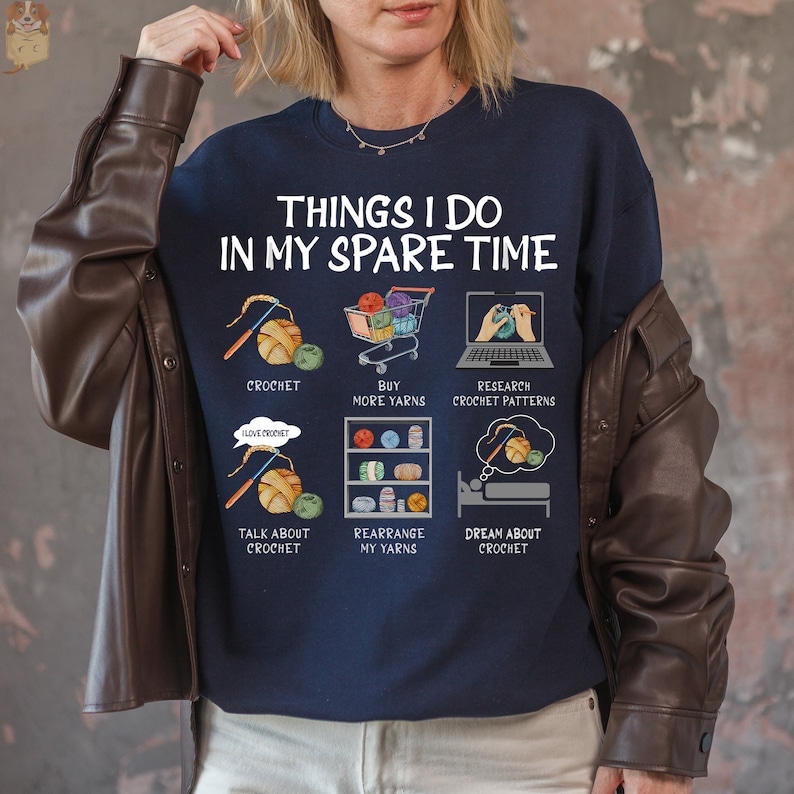May include: A navy blue sweatshirt with the text "Things I do in my spare time" and illustrations of yarn, crochet hooks, and shopping carts. The illustrations depict activities related to crocheting, such as buying yarn, researching patterns, and dreaming about crochet.