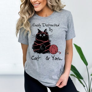 May include: Gray t-shirt with a black cat tangled in red yarn and a red ball of yarn. The text on the shirt reads "Easily Distracted By Cat & Yarn".
