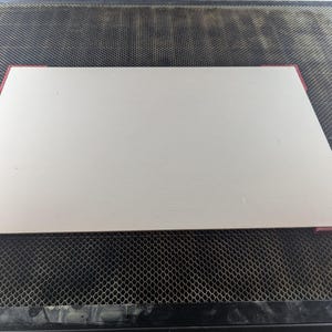 May include: A white rectangular cutting board with red edging. The board is on a black mesh surface.