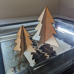 May include: Two wooden Christmas tree ornaments, one larger than the other, are sitting on a black surface. The trees are made of plywood and have a natural wood finish.